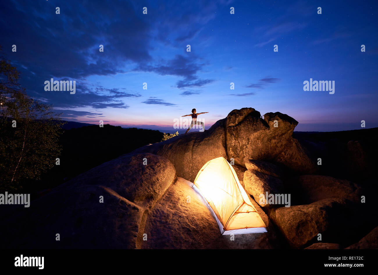 Il campeggio sulla formazione di roccia. Luminose tenda turistica e silhouette di slim ragazza turistica facendo esercizi yoga sulla cima della montagna contro il cielo blu scuro al tramonto. Sport, turismo concept. Virabhadrasana Foto Stock