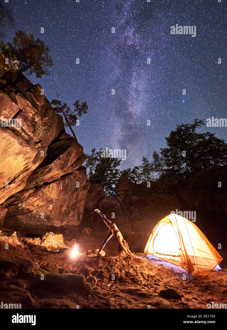 Campeggio in mezzo a un enorme roccia ripida formazioni. Illuminato da campfire donna facendo esercizi yoga vicino al piccolo turista tenda su una chiara notte cielo stellato sfondo. Uno stile di vita sano concetto. Adho Mukha Shvanasana Foto Stock