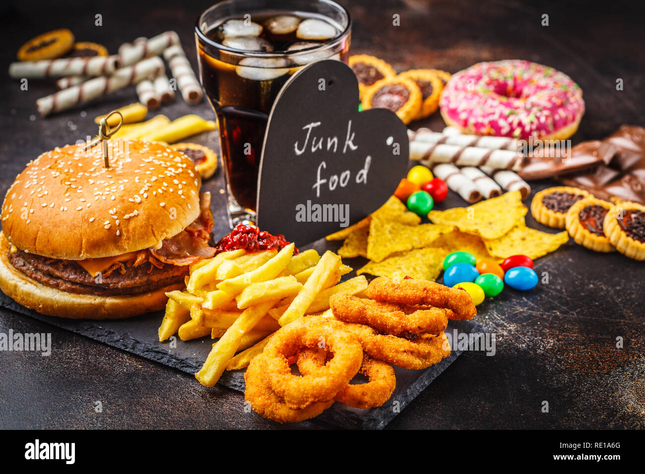 Junk food concept. Cibo malsano sfondo. Fast food e zucchero. Burger, dolci, patatine, cioccolato, ciambelle, soda su uno sfondo scuro. Foto Stock