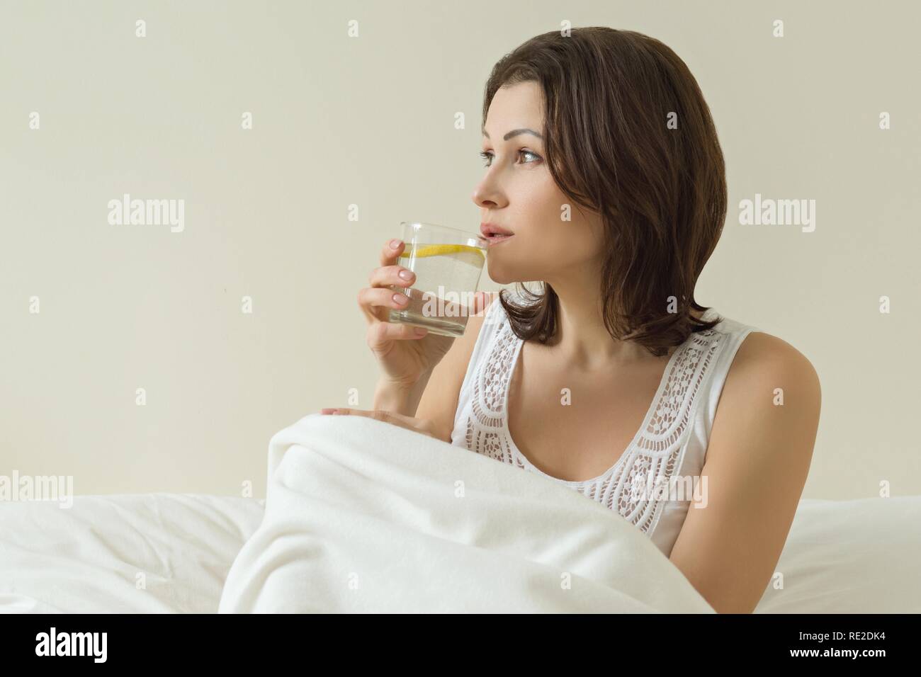 Donna di mezza età gode di acqua con un limone seduto nel letto. La prima mattina bere, bevanda antiossidante. Foto Stock