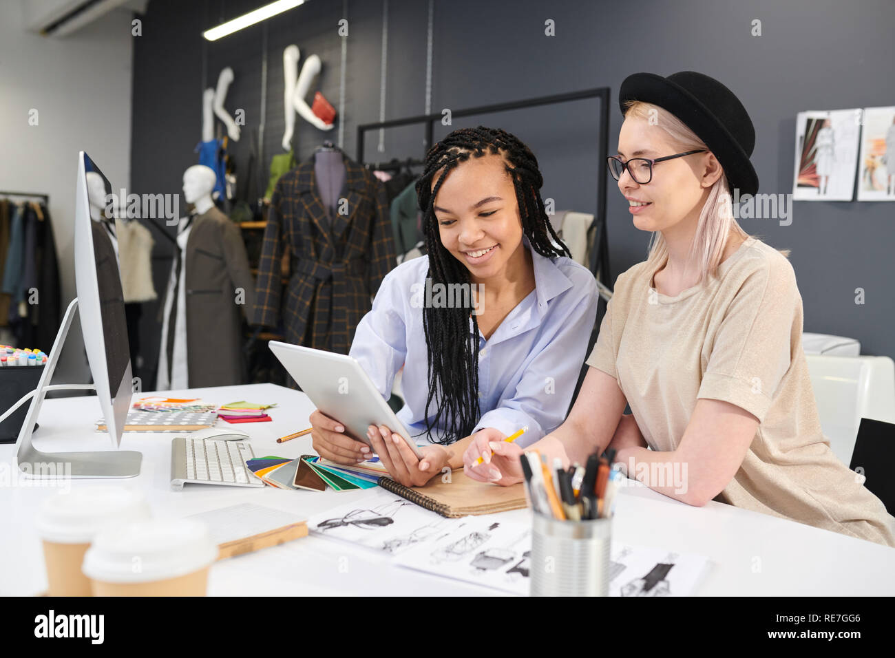 Designer di moda seduti a tavola tra schizzi e tavolozze e con tavoletta digitale insieme in officina Foto Stock