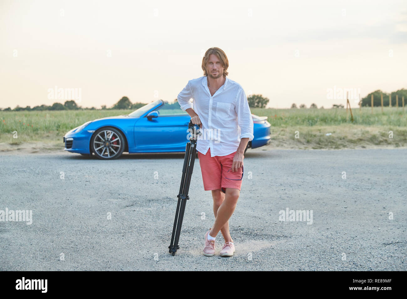 La bellissima giovane uomo brutale sorge in corrispondenza di un blu auto sportiva al tramonto, egli è vestito con una camicia bianca con maniche corte e pantaloncini rossi Foto Stock