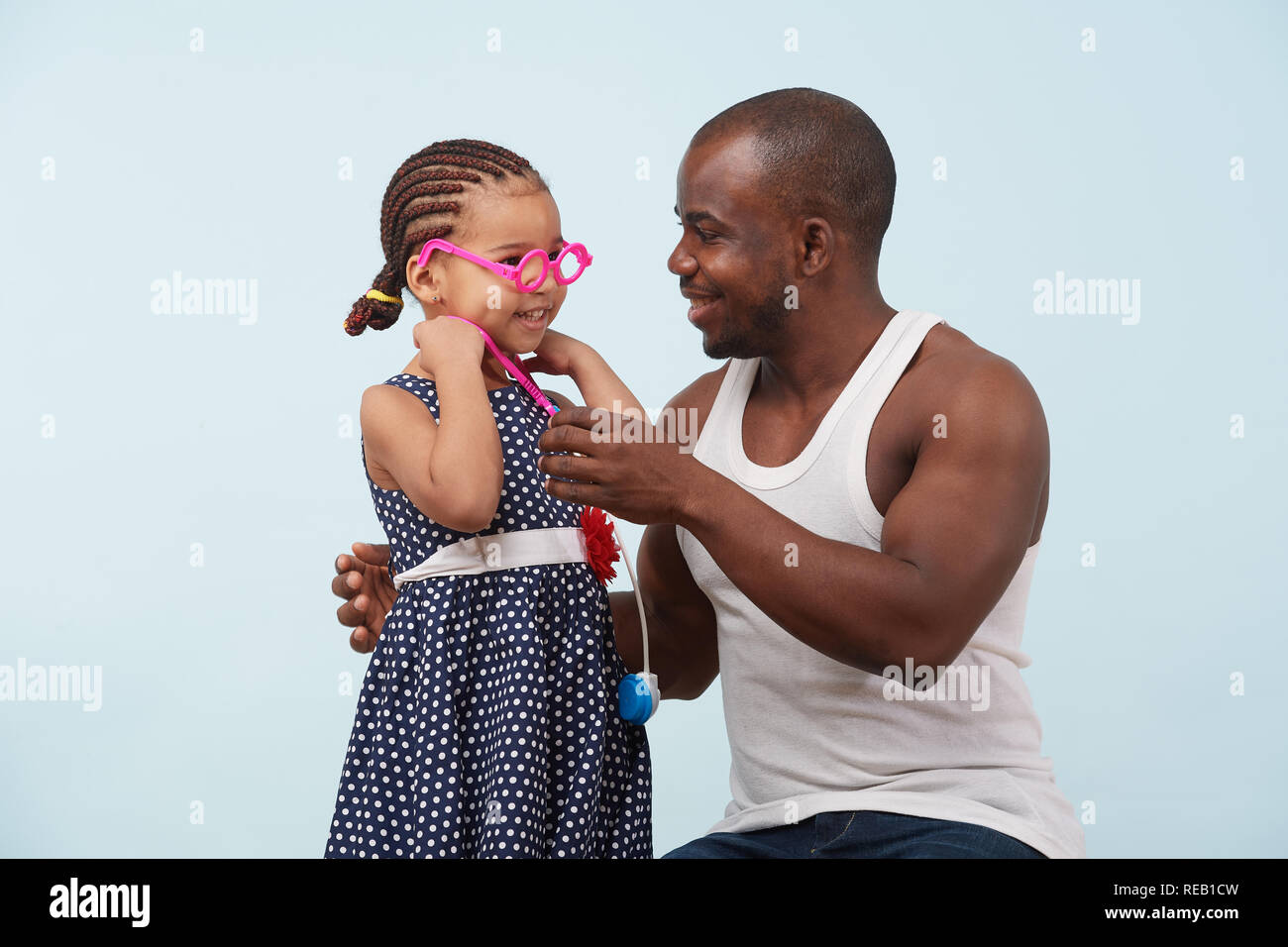 Felice padre e figlia riproduzione medico con stetoscopio giocattolo contro il fondo azzurro. Indossa blue polka dot dress e trecce. Egli è in grassetto, ho Foto Stock