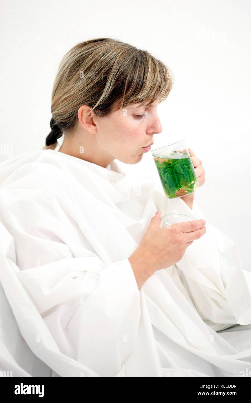 Una giovane donna con un mal di gola e freddo nel letto, con una sciarpa di bere il tè alla menta con foglie di menta fresca e rilassante Foto Stock