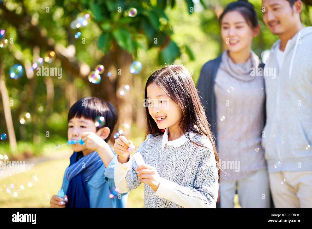 Poco ragazzi asiatici un ragazzo e una ragazza sorella e fratello soffiare bolle in un parco con i genitori guardando da dietro. Foto Stock