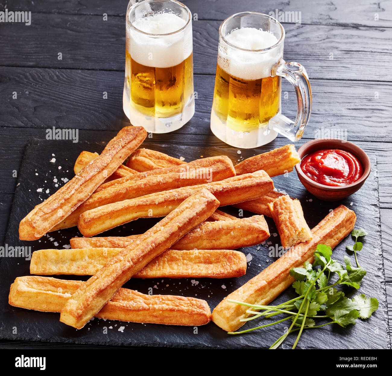 Close-up di formaggio cannucce, Cheesy Grissini su un taglio di legno con salsa di pomodoro e due bicchieri di vetro espanso con birra fredda su di un legno nero Foto Stock