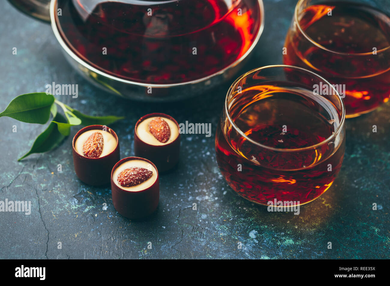 Due bicchieri di vetro e teiera con il tè nero e cioccolatini su sfondo scuro. Foto Stock