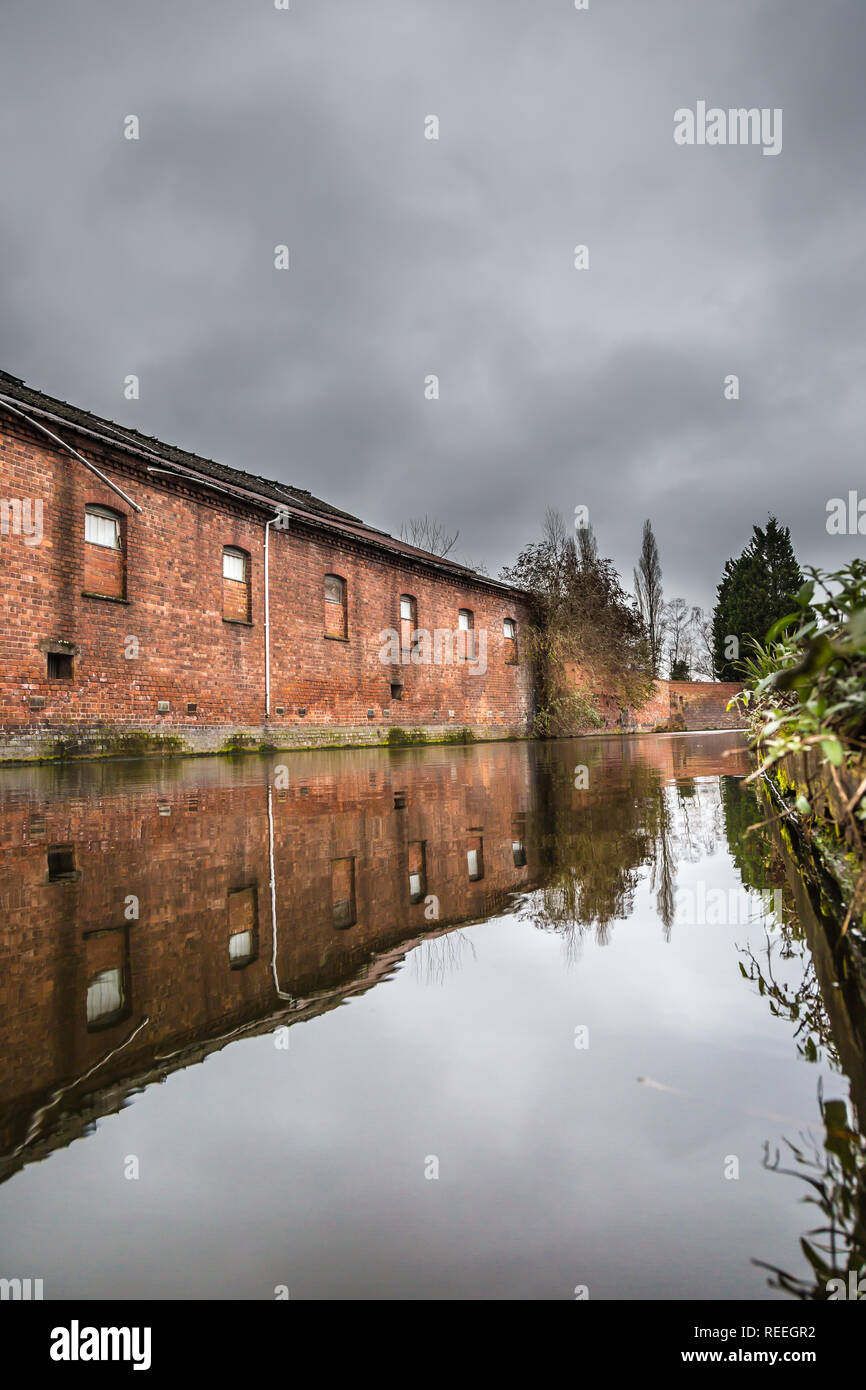 Arty, ritratto di vecchia, dismesse, moquette edificio Mill si riflette ancora in acqua del canale a Kidderminster, Regno Unito su un inverno mattina con il grigio moody sky. Foto Stock