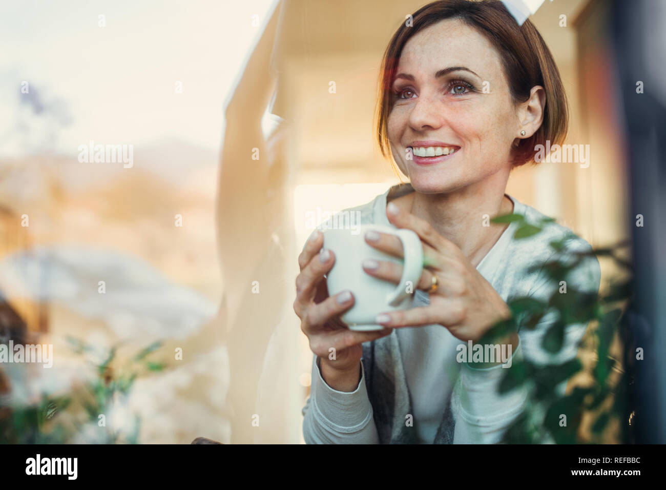 Una giovane donna con la tazza di caffè guardando fuori della finestra. Shot attraverso il vetro. Foto Stock