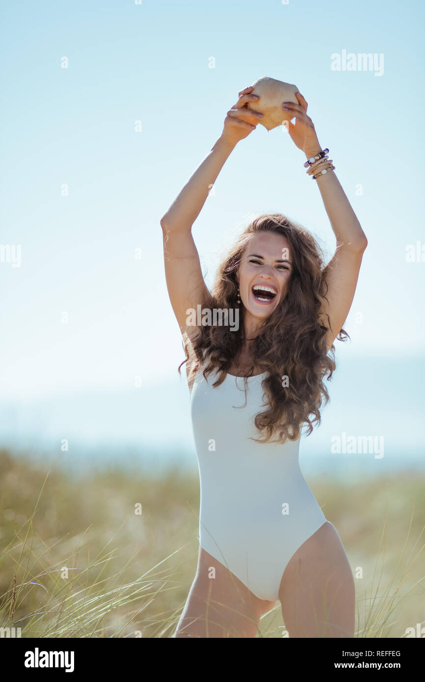 Sorridente giovane donna in bianco beachwear con noce di cocco sulla spiaggia. Noce di cocco utilizzata nell'industria cosmetica per migliorare la salute e l'aspetto della sk Foto Stock
