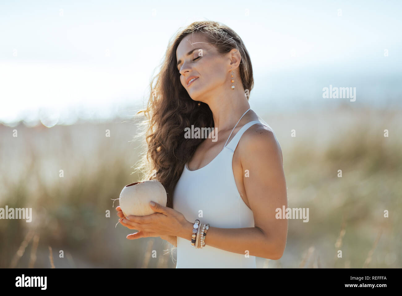 Moderno e rilassato donna in bianco beachwear sulla costa dell'oceano holding di noce di cocco. Il Cocco può aiutare a ottenere brillanti e capelli setosi. spiaggia vocazione. wild beach w Foto Stock