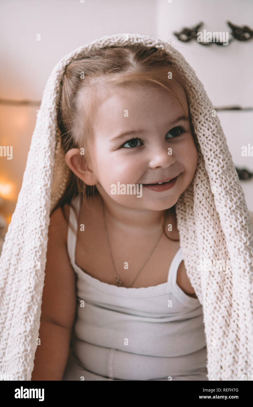 Grazioso piccolo ragazza sorridente coperto in un grande plaid o sciarpa a casa Foto Stock