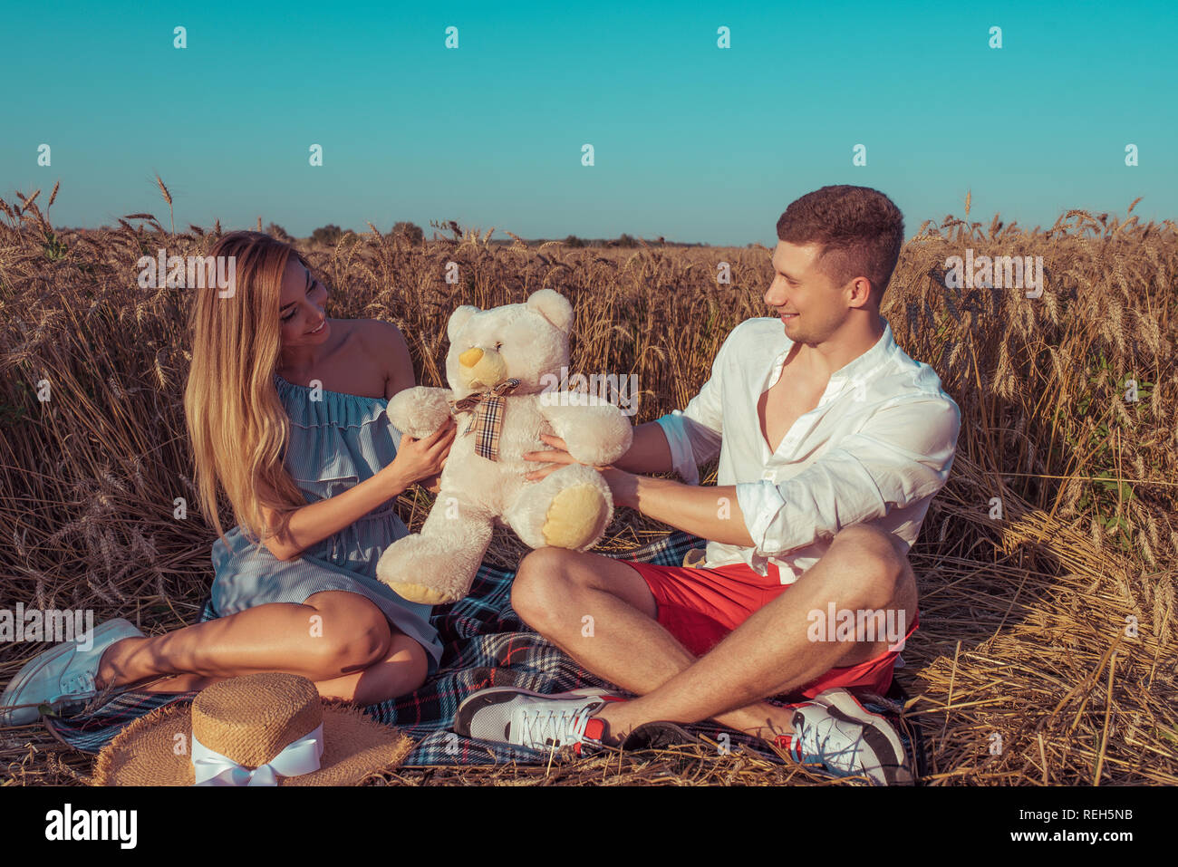 Una ragazza con i capelli lunghi riceve orsacchiotto dono dalla sua giovane ragazzo. Sorridenti rallegratevi. Seduto in un campo di grano in estate. Data romantico, sposi novelli, felice nuova famiglia. Foto Stock