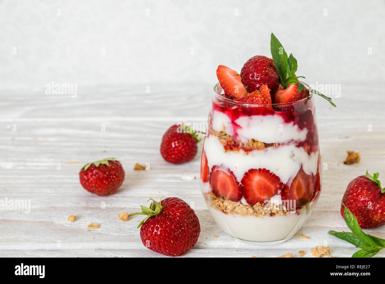 Yogurt alla fragola parfait con muesli, menta e frutti di bosco freschi in un vetro bianco su un tavolo di legno. una sana prima colazione Foto Stock