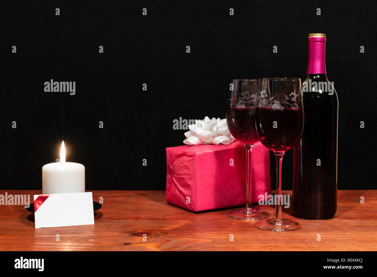 Bella inciso bicchieri di vino e una bottiglia di vino rosso, bianco candela, avvolti presente con arco sul tavolo di legno con il nome di tag su sfondo scuro. Foto Stock