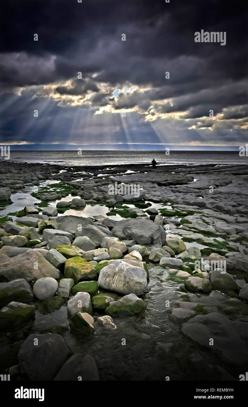 Spiaggia rocciosa con cielo tempestoso e luce spettacolare e una persona in silhouette a distanza dal concetto di solitudine bordo dell'acqua Foto Stock