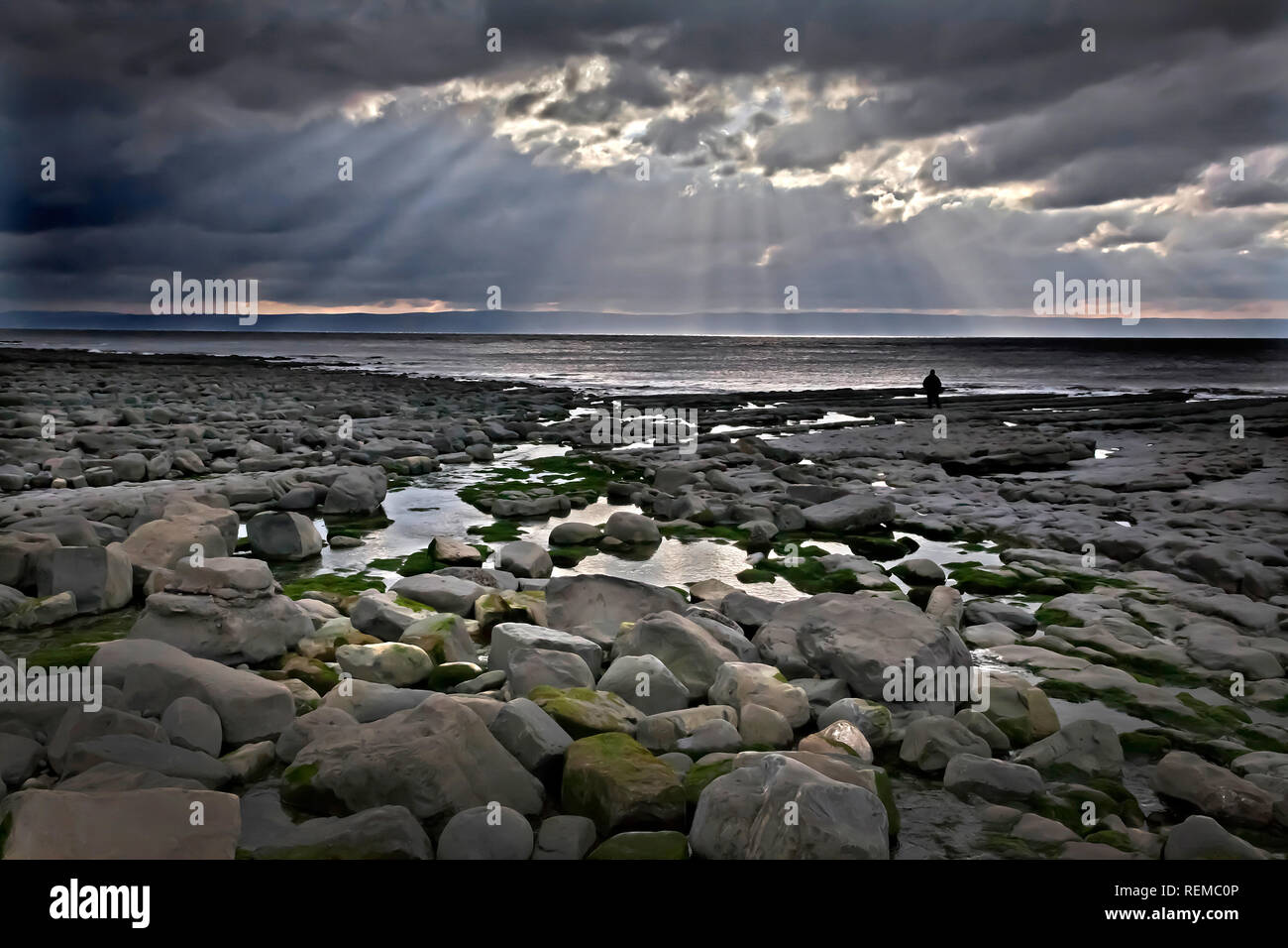 Spiaggia rocciosa con cielo tempestoso e luce spettacolare e una persona in silhouette a distanza dal concetto di solitudine bordo dell'acqua Foto Stock