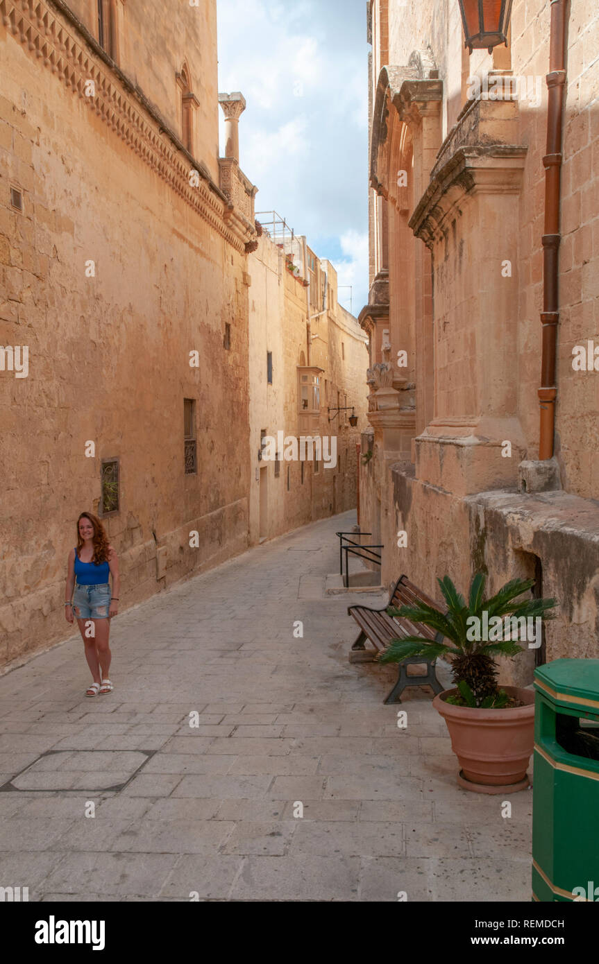 Giovane donna a piedi attraverso le tipiche strade strette tra case di calcare nell'antica città fortificata di Mdina a Malta. Foto Stock