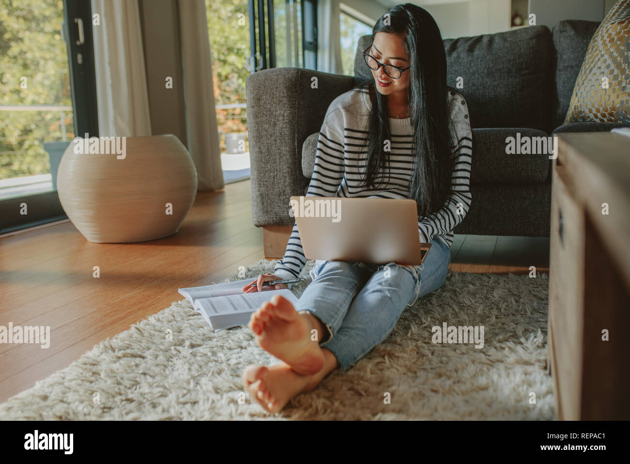 Donna seduta sul pavimento appoggiato al divano guardando il libro con il computer portatile sul suo grembo. Studentessa studiare a casa. Foto Stock