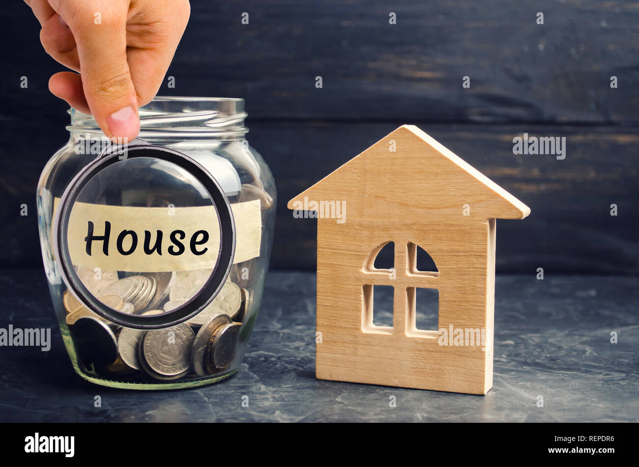 Un vaso di vetro con monete e una casa in legno e la scritta 'casa'. L'accumulo di denaro per comprare una casa. Affitta un appartamento. Acquisto di una casa. Foto Stock