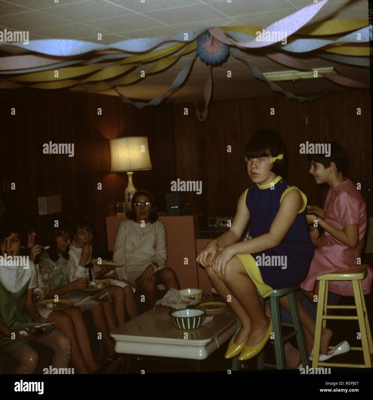 Vista interna di un gruppo di giovani adolescenti di sesso femminile in decorate salotto di una casa suburbana durante una festa, seduti insieme con un aspetto imbarazzante, uno teen seduto su un alto sgabello in primo piano, 1965. () Foto Stock