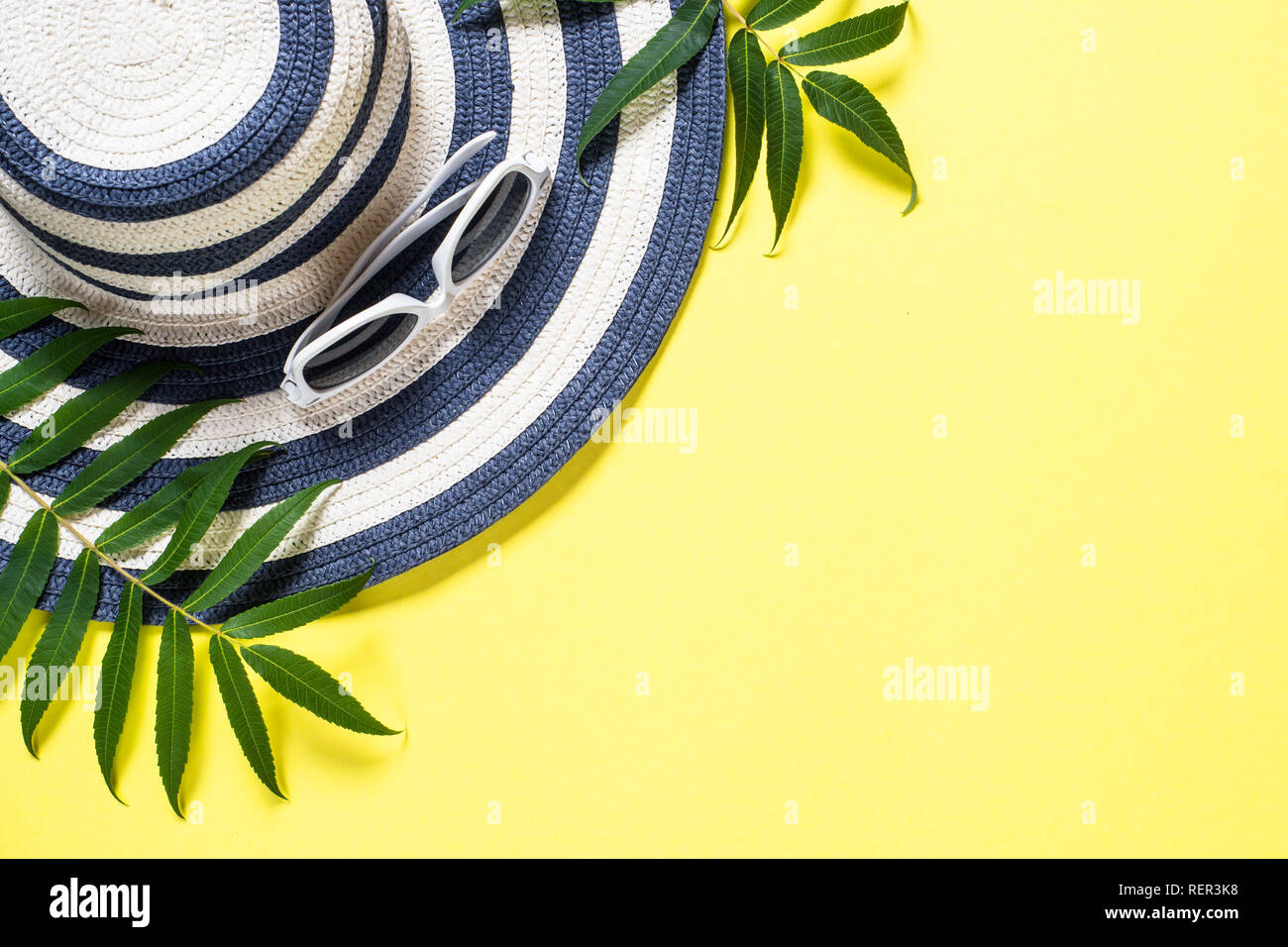 Striped cappello, occhiali da sole e foglie tropicali su sfondo giallo. Estate vacanze viaggi sfondo. Vista da sopra con copia spazio. Foto Stock
