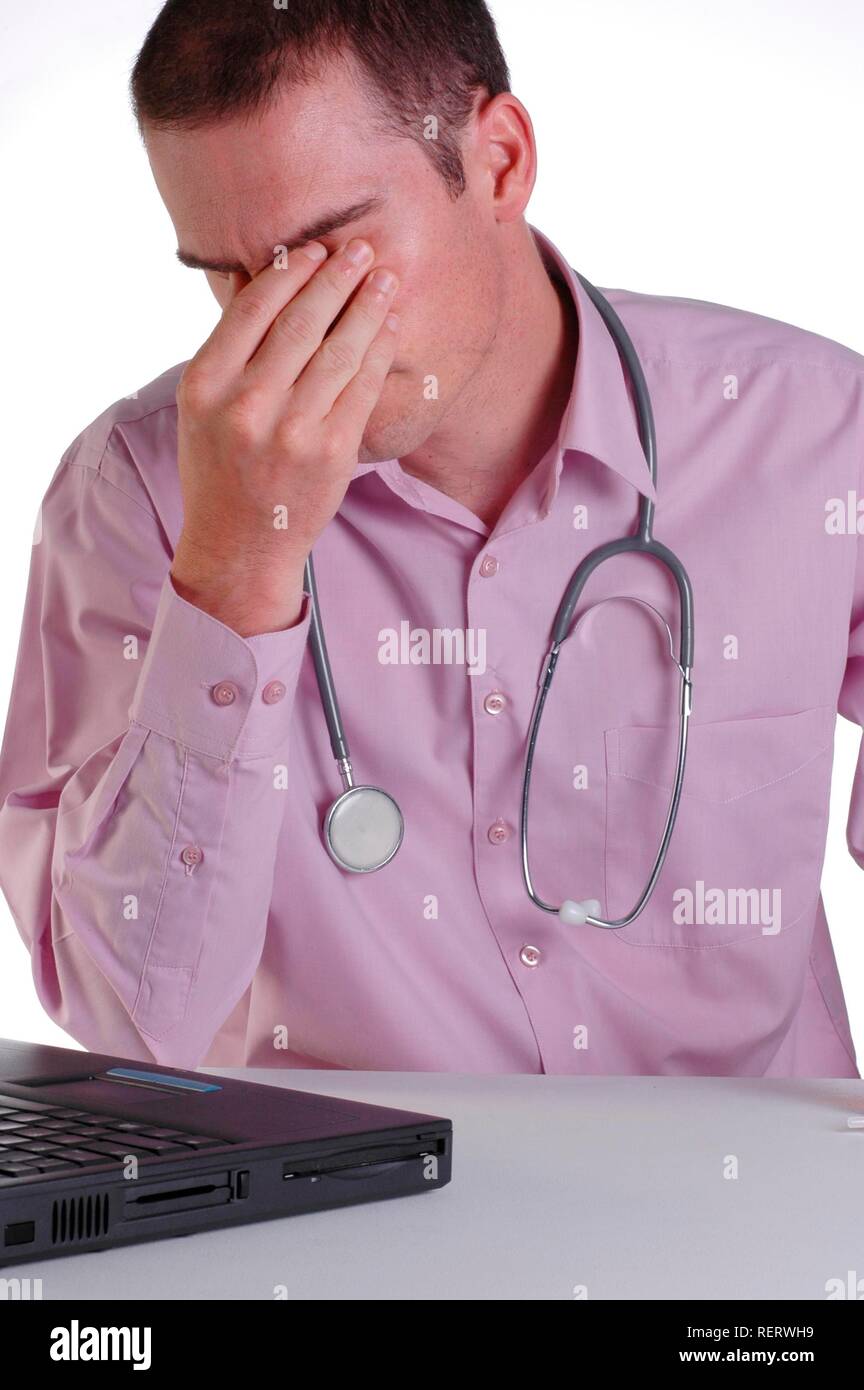Un giovane maschio medico al suo banco stropicciandosi gli occhi con fatica da orari di lavoro eccessivamente prolungati Foto Stock