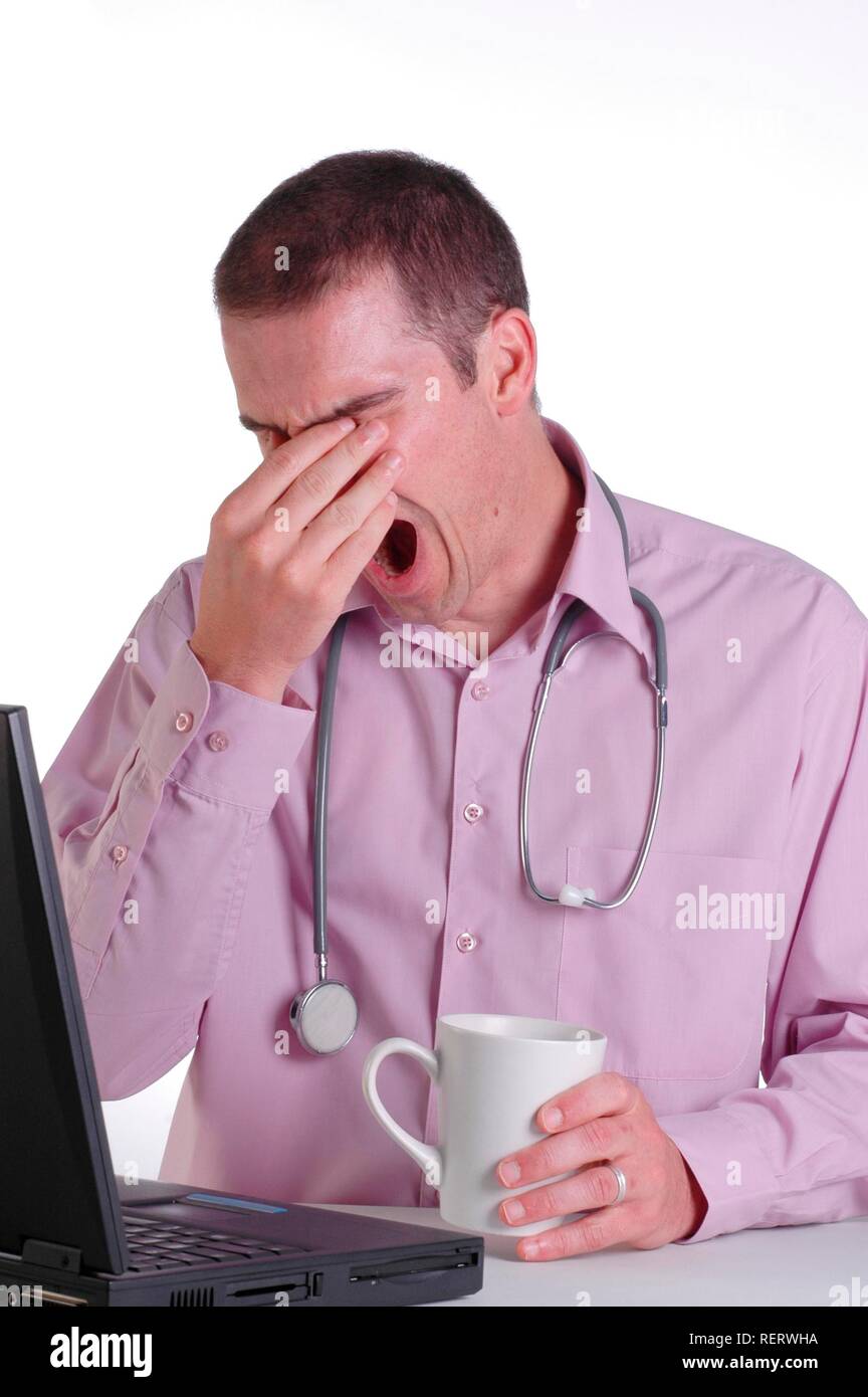 Un giovane maschio medico al suo banco strofinando gli occhi e sbadigli con fatica da orari di lavoro eccessivamente prolungati Foto Stock