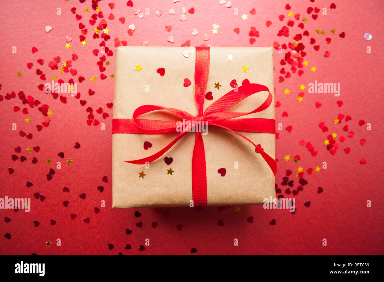 Confezione regalo avvolto in colore marrone carta craft e legato con fiocco rosso scuro su sfondo rosso con red coriandoli. Foto Stock