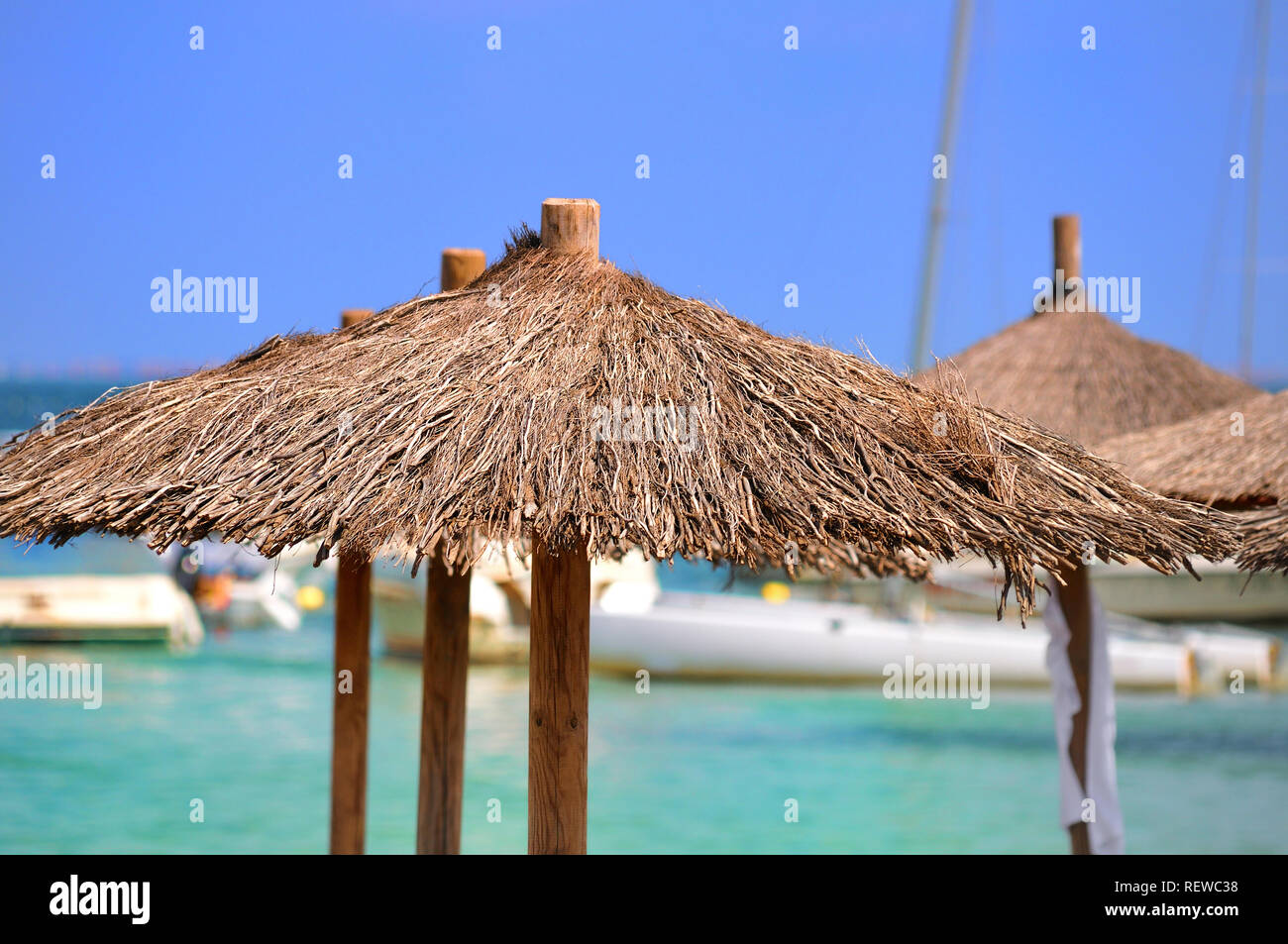 Barche ormeggiate galleggiante in cristalline acque turchesi dietro il rustico parasol ombrelloni. Una visione di un sogno spiaggia vacanza. Foto Stock
