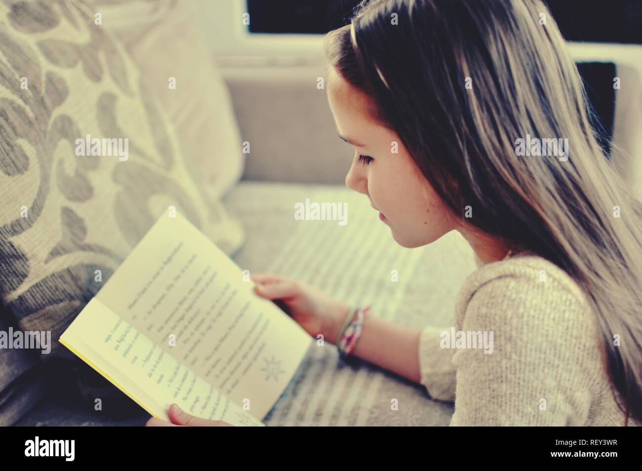 Ritratto di una bionda caucasico bianco 9 anni ragazza seduta vicino ad un divano e la lettura di un libro. Il bambino ha i capelli lunghi. Vista frontale, il fuoco selettivo. Foto Stock