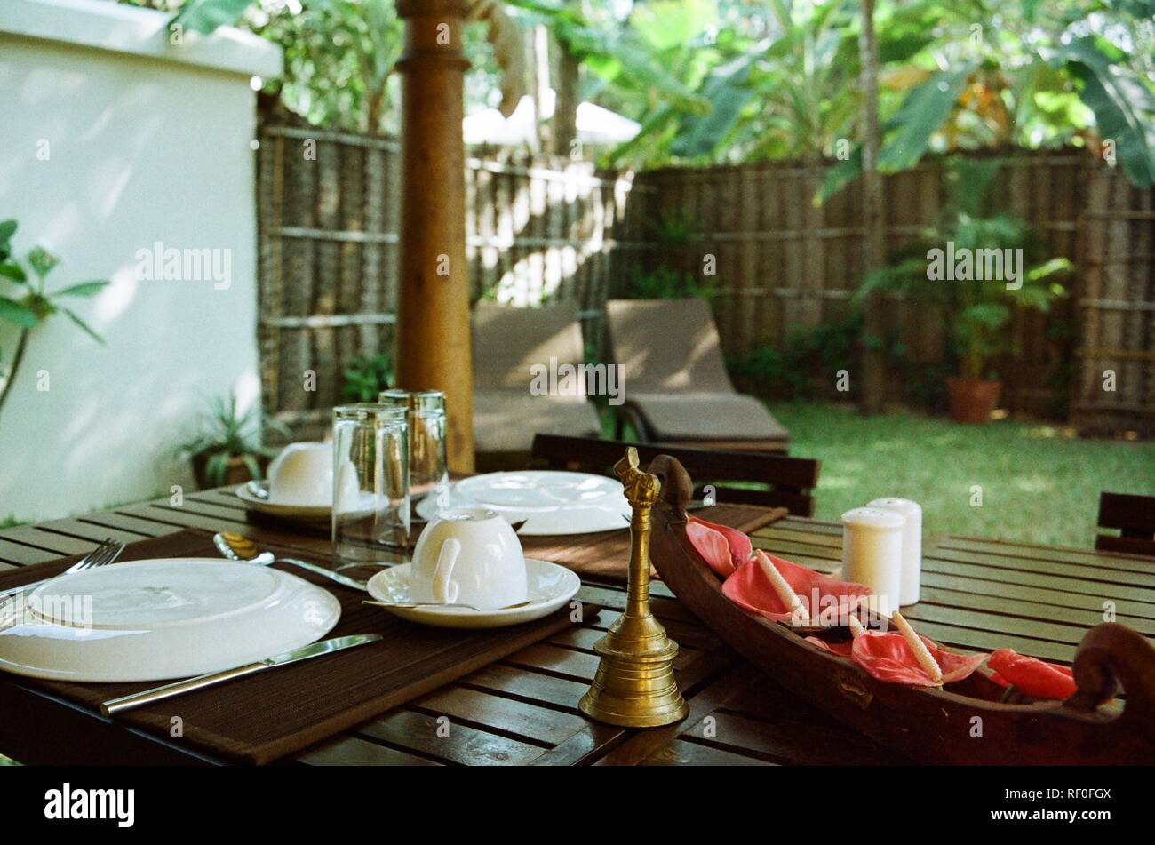 Una fresca di cui tavolo con fiori e una campana di ottone nel giardino privato di un convertito casa di pescatori in Alappuzha, Kerala, India. Foto Stock
