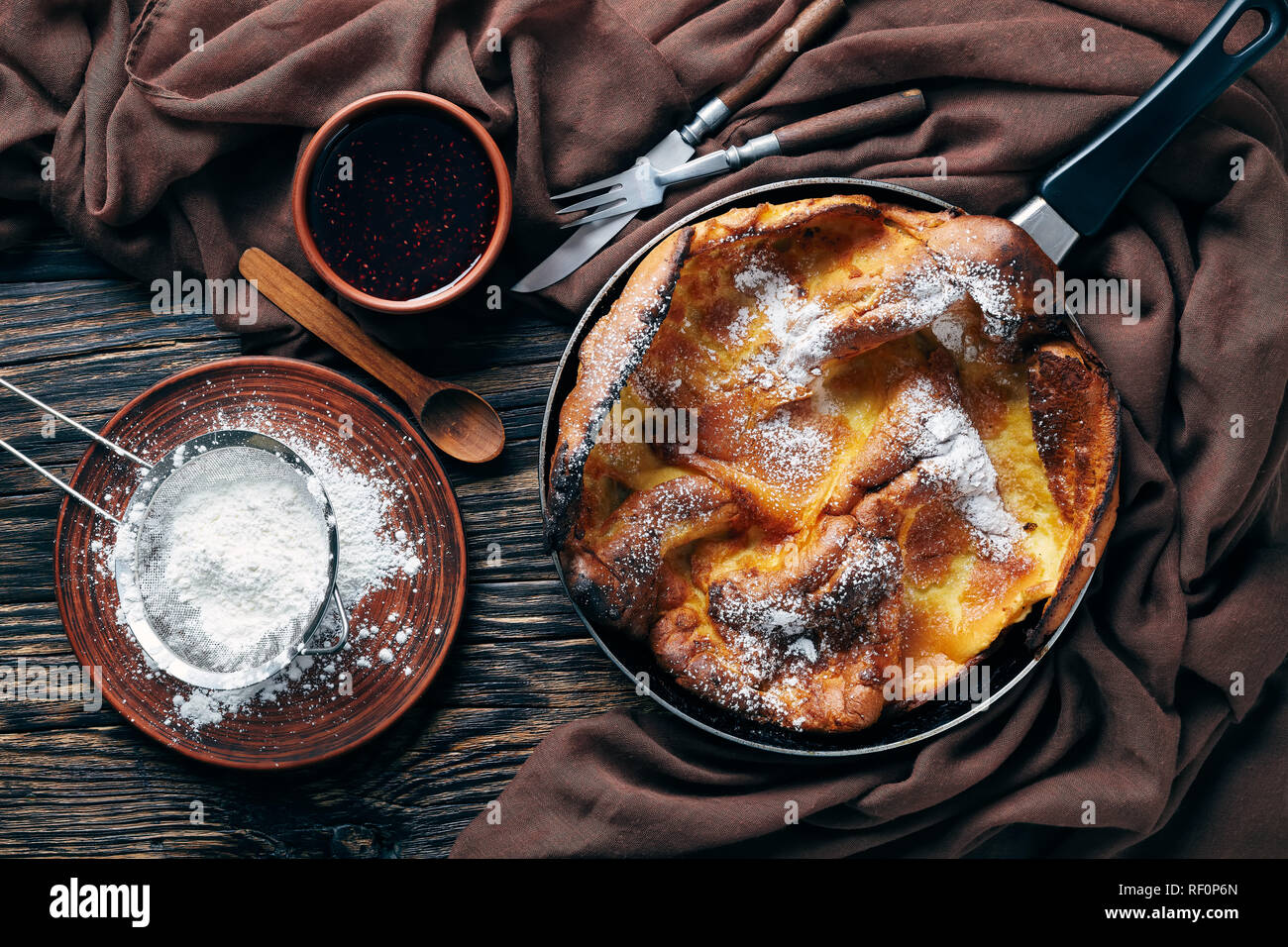 Dutch baby pancake spolverati con zucchero a velo in una padella con confettura di lamponi e panno marrone su un vecchio tavolo rustico, ricetta classica, vista da abo Foto Stock