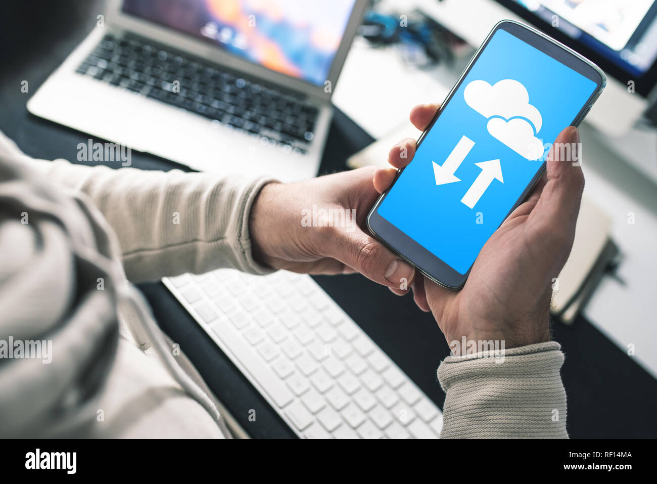 Uomo seduto alla scrivania in ufficio utilizzando il servizio cloud sullo smartphone per il trasferimento e la sincronizzazione dei dati Foto Stock