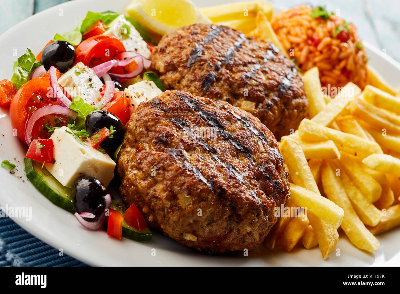 Pan-sparò bifteki o frikadelle con insalata greca, pomodoro riso pilaf e patatine fritte in una vista ravvicinata su una piastra Foto Stock