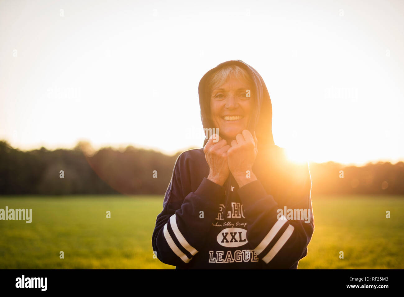 Ritratto di sorridere senior donna che indossa una felpa con cappuccio in piedi sul prato rurale al tramonto Foto Stock