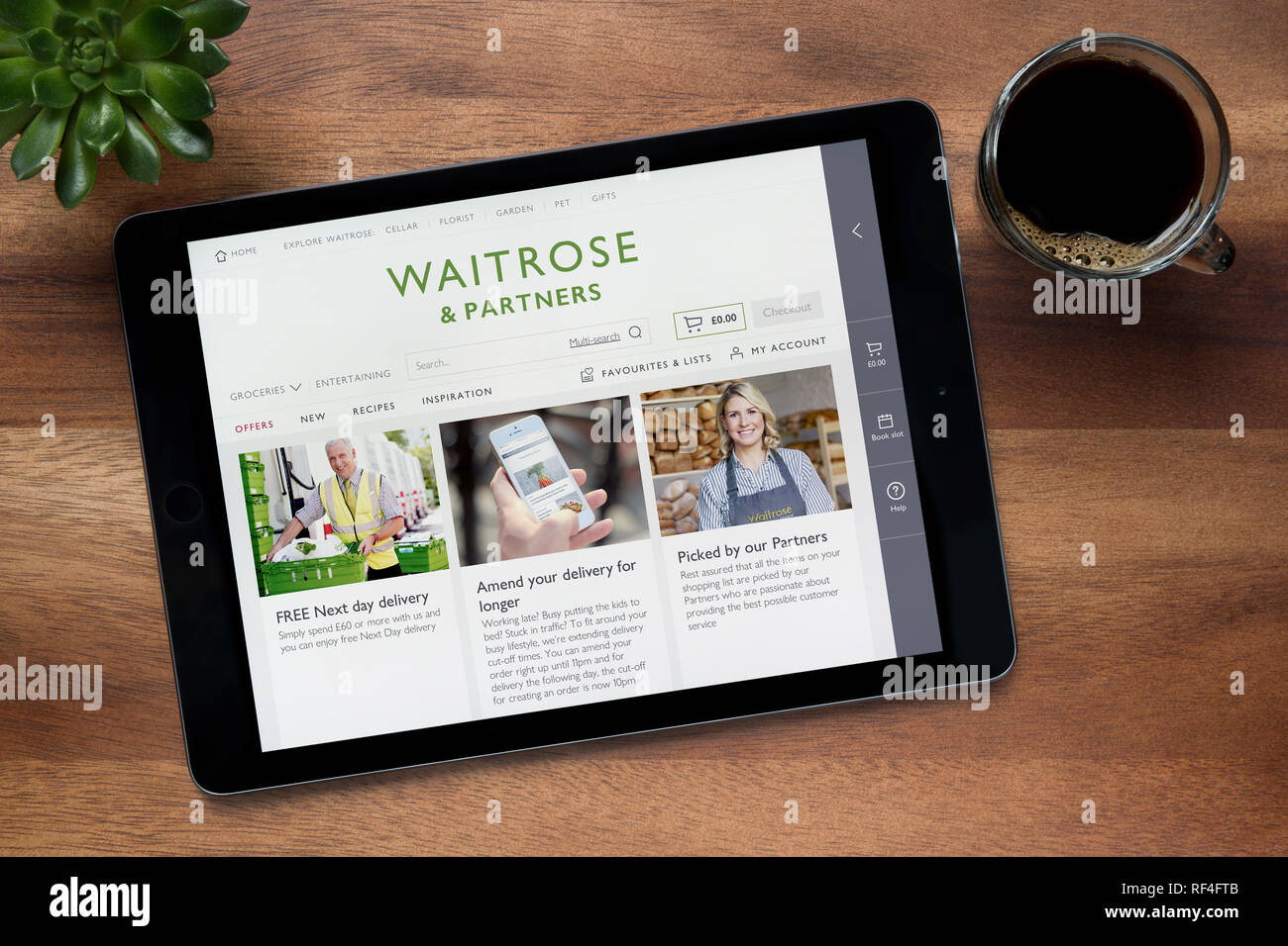 Il sito web di Waitrose e partner è visto su un tablet iPad, su di un tavolo di legno lungo con un caffè espresso e una pianta di casa (solo uso editoriale) Foto Stock