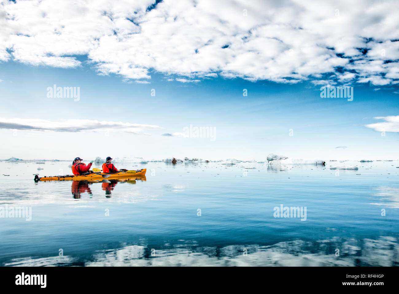 Klerckbukta Nordaustlandet Svalbard | NORDAUSTLANDET, Svalbard — Due kayakisti con giacche rosse remano un tandem giallo su acque calme come uno specchio, punteggiate di ghiaccio galleggiante, con la lontana fronte di ghiaccio di Bråsvellbreen visibile all'orizzonte. Questo sbocco del ghiacciaio Austfonna incontra il mare lungo la remota costa sudorientale di Nordaustlandet, a portata di Klerckbukta. Foto Stock