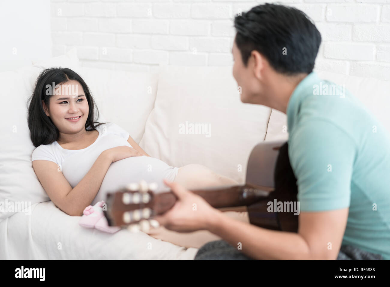 Bella asiatica donna incinta e Asian bel marito sono sorridente e suonare la chitarra acustica sul letto di casa. Foto Stock