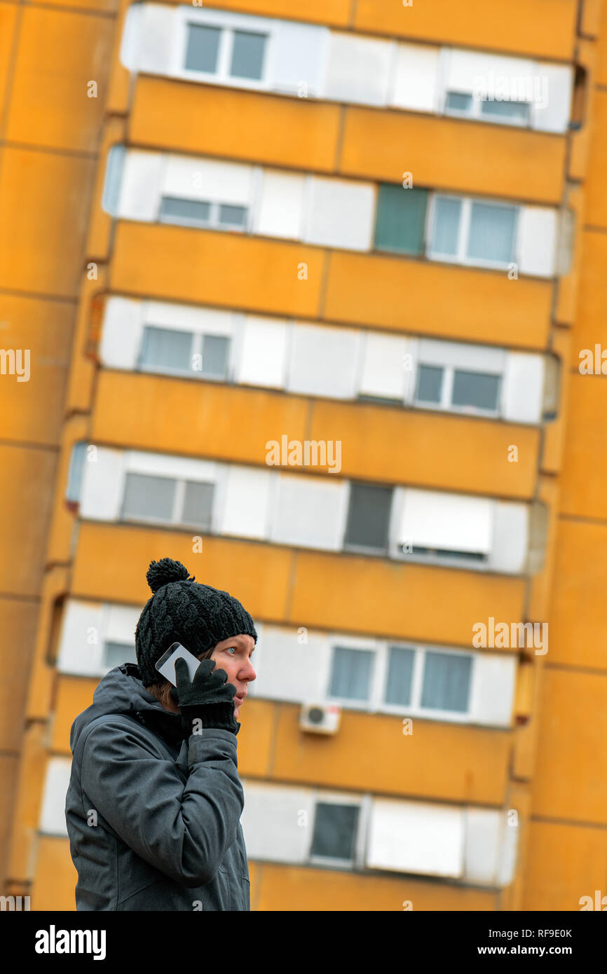 Donna che parla al telefono cellulare in strada in inverno. Grave femmina caucasica durante la conversazione telefonica con il brutto edificio di appartamenti in background. Foto Stock