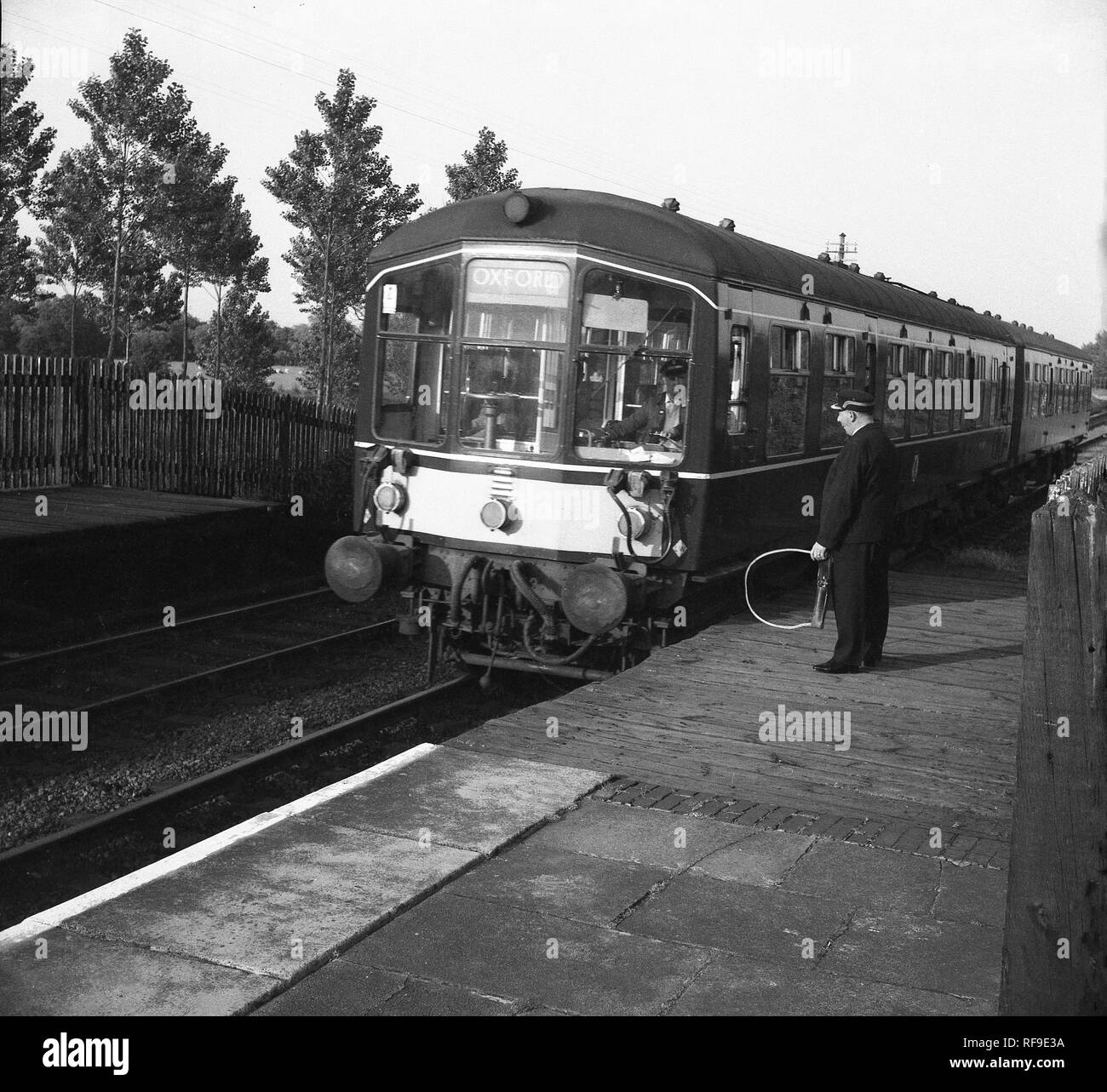 Settanta i treni diesel provenienti in una stazione ferroviaria con una stazione di attesa guard in piedi sul legno parte anteriore platform tenendo un attrezzo metallico, Inghilterra, Regno Unito. Foto Stock