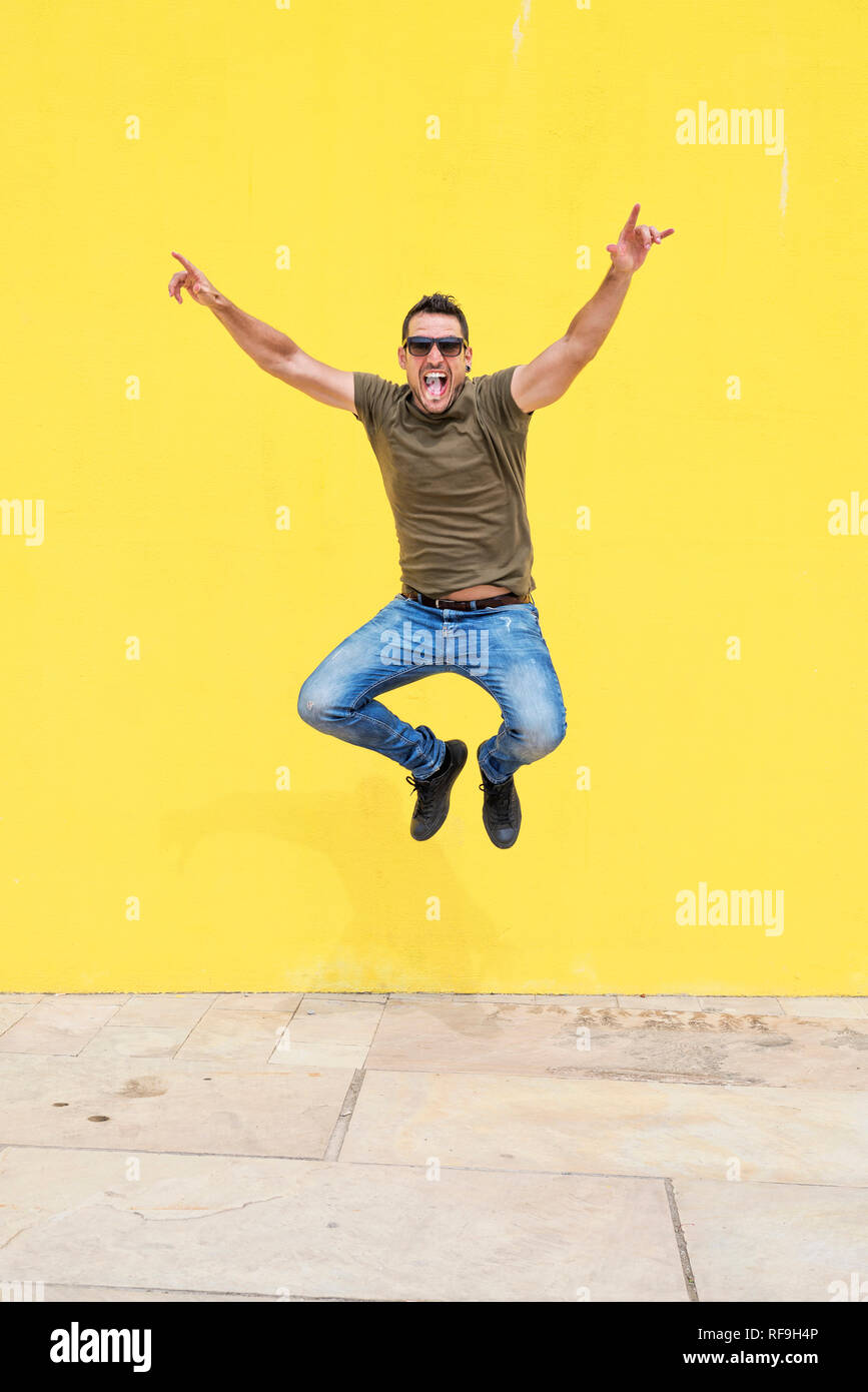 Vista frontale di un giovane uomo felice indossando occhiali da sole jumping contro un luminoso giallo parete in una giornata di sole Foto Stock