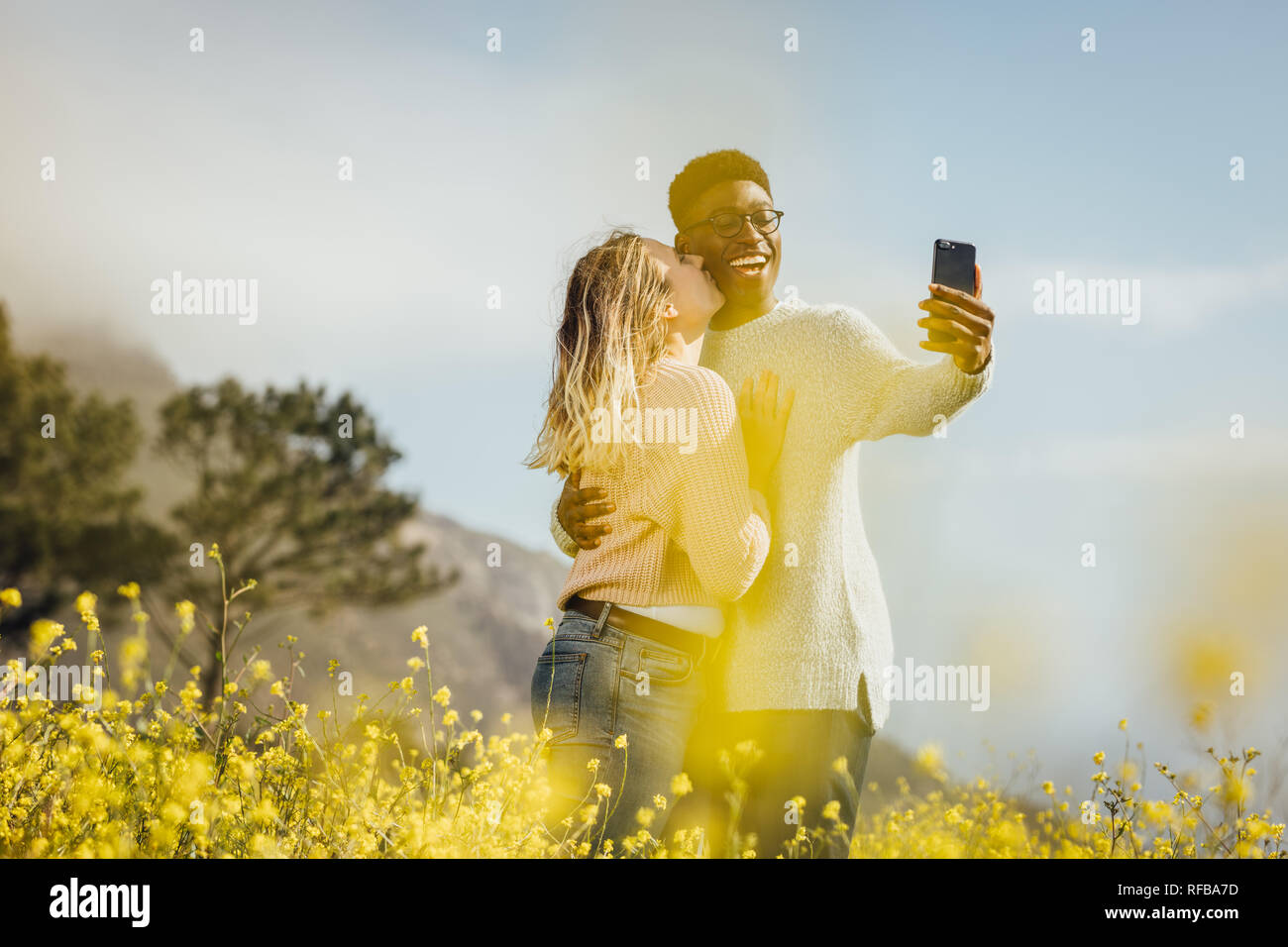 Coppia romantica in piedi all'aperto tenuto selfie. L'uomo africano tenendo selfie sul cellulare durante la donna baciare lui sulla sua guancia. Foto Stock