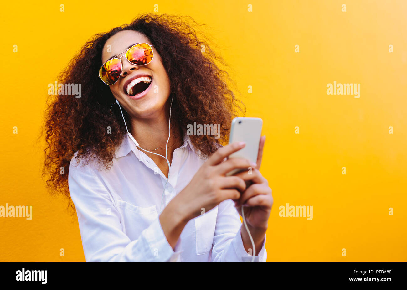 Ridendo giovane donna ascoltando musica in cuffia utilizzando un telefono cellulare. Ragazza africana con capelli ricci indossando occhiali da sole contro la parete gialla in ascolto Foto Stock