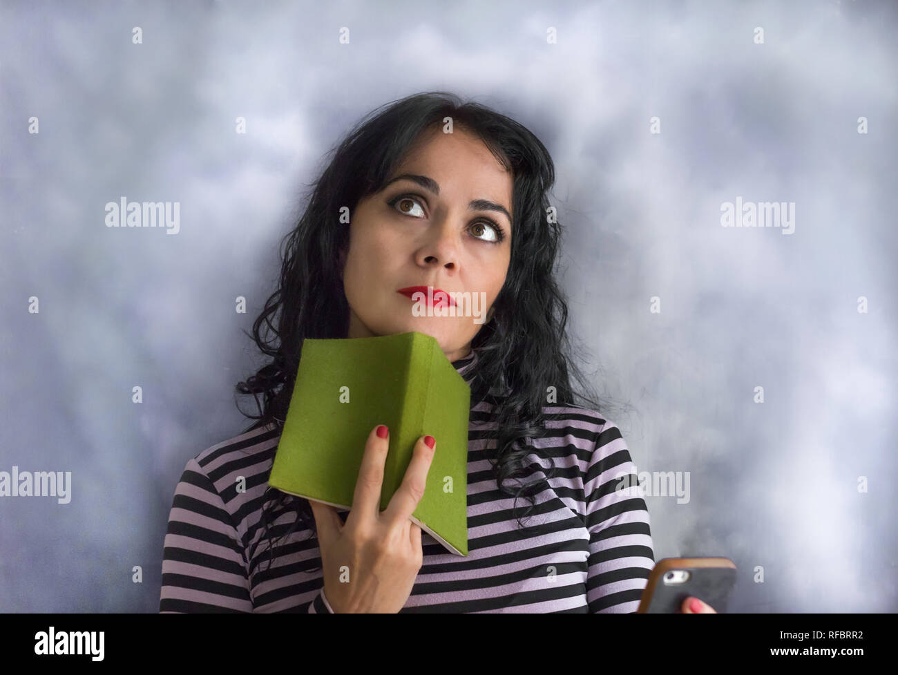 Bruna giovane donna con il maglione a strisce con un libro sul suo mento pensando ad una questione, isolata su uno sfondo grigio Foto Stock