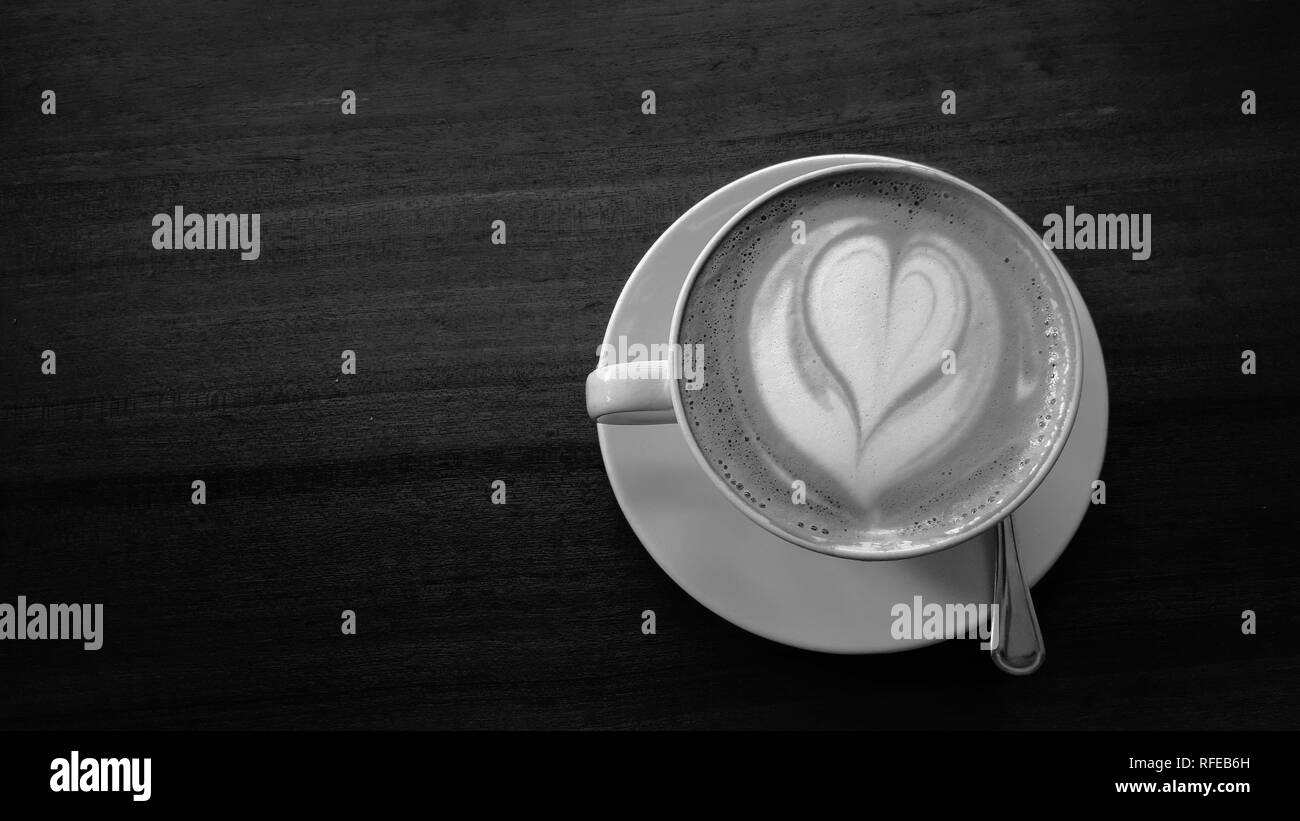 Una foto in bianco e nero di un bianco caffè e piattino pieno di caffè e ha un cuore schiuma latte art sulla parte superiore e seduti su un tavolo di legno. Vista dall'alto. Foto Stock