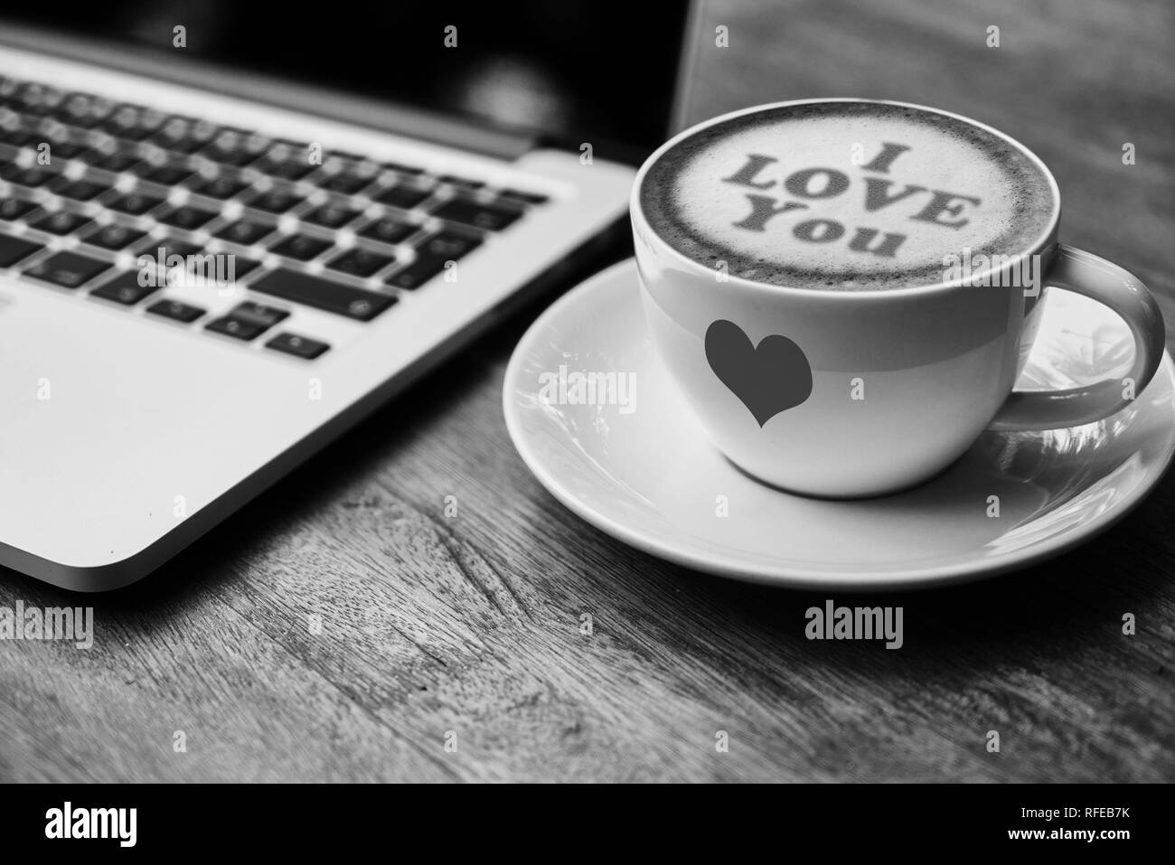 Una foto in bianco e nero di un bianco tazza da caffè con piattino con una schiuma latte art che dice "ti amo" e seduto accanto a un computer portatile su un tavolo di legno. Foto Stock
