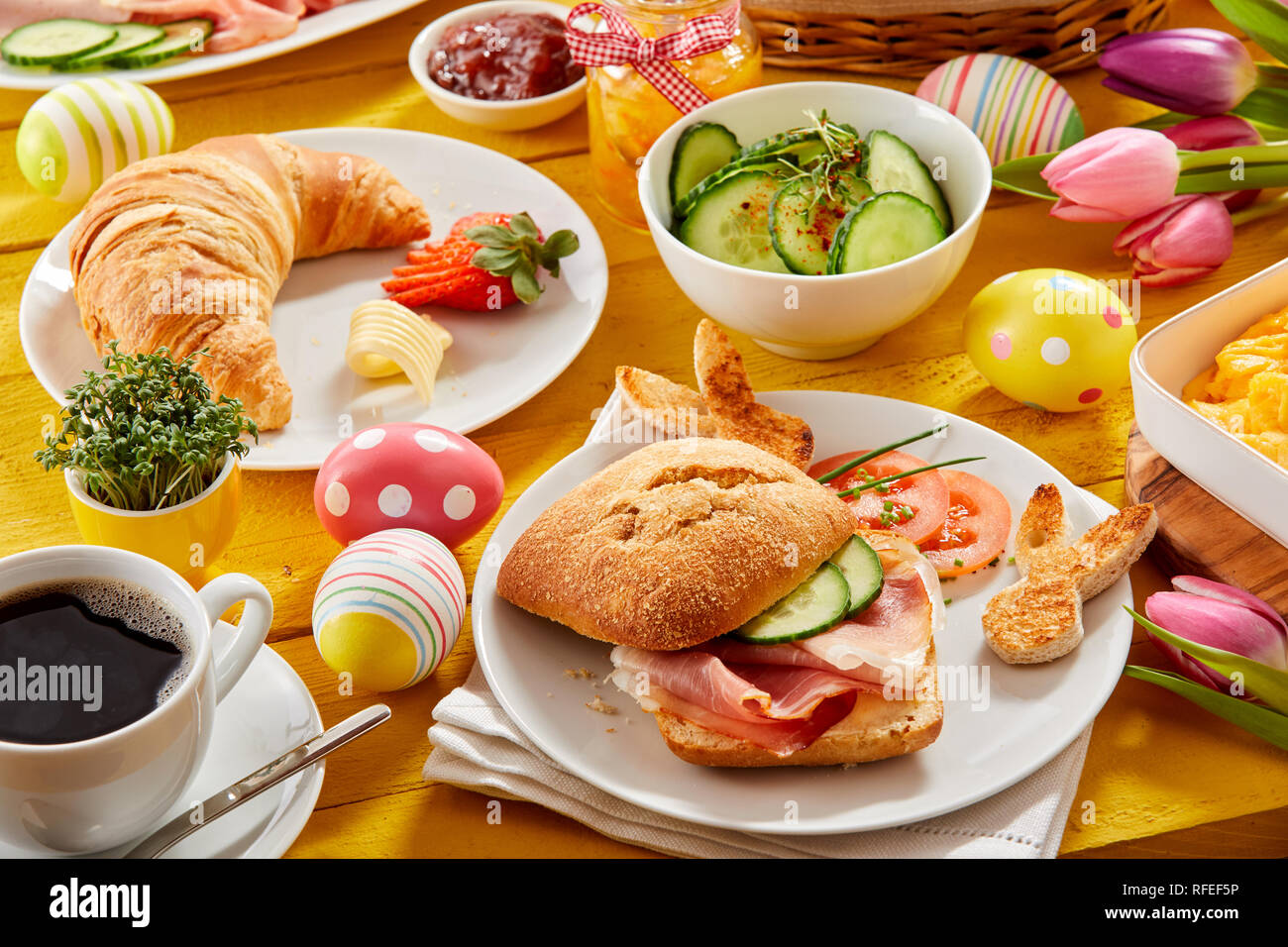 Gustoso brunch pasquale o in primavera prima colazione con croissant fresco, prosciutto o pancetta rotolo e caffè su una tavola decorata con colorate le uova di Pasqua e tulipani Foto Stock