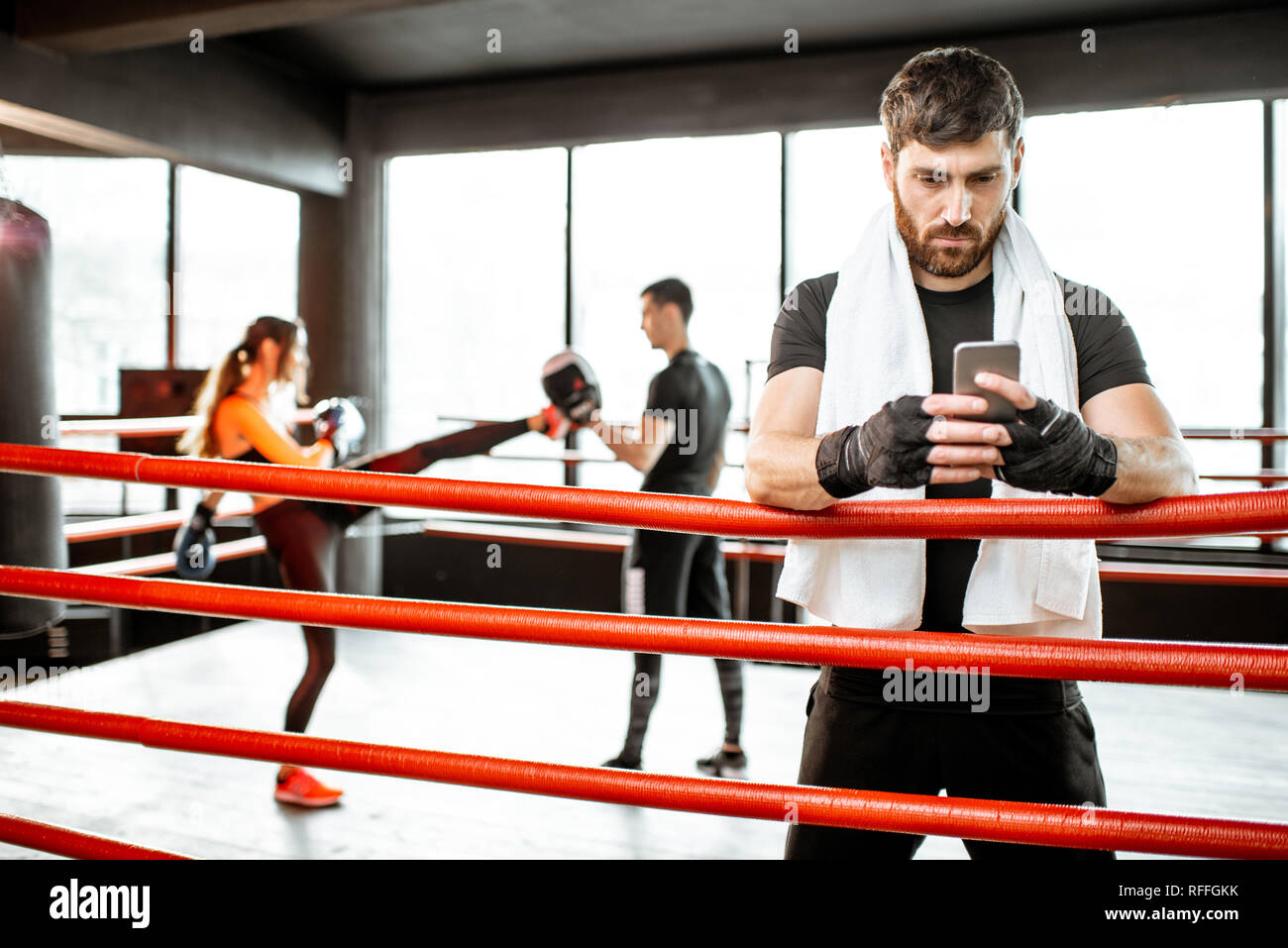 Uomo appoggiato con il telefono dopo la formazione su pugilato ring con persone boxe sullo sfondo in palestra Foto Stock