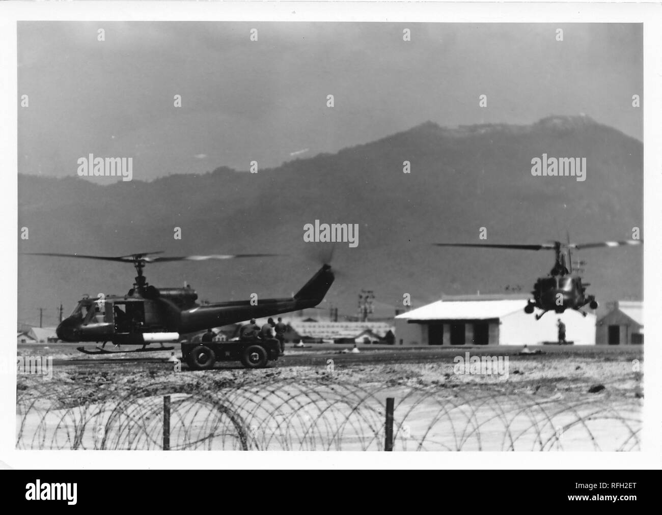 Fotografia in bianco e nero, che mostra due Bell UH-1 Iroquois ("Huey") elicotteri, sia di atterraggio o di decollo da una base militare, con diversi uomini in un aperto Jeep e caserme o edifici amministrativi visibile sullo sfondo, fotografato durante la Guerra del Vietnam, 1968. () Foto Stock